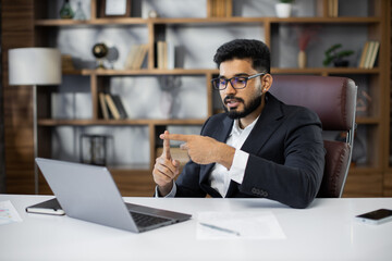 Young bearded businessman using laptop computer during video call working in office. Concentrated adult successful man wearing official suit sitting at wooden desk indoor.