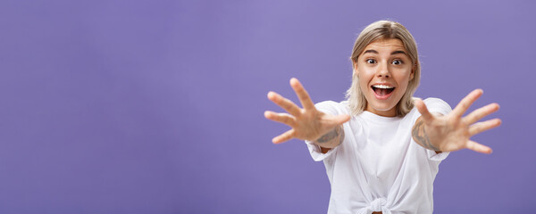 Waist-up shot of amused and excited attractive stylish young woman in white t-shirt pulling hands at camera with desire smiling thrilled and happy wanting hug or take something over purple background