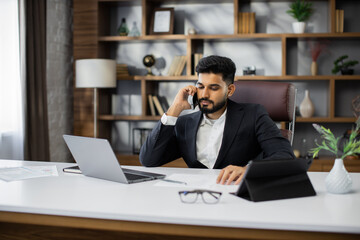 Successful young entrepreneur having phone consultation with business partners, sitting at desk in modern office. Attractive businessman focused on work issues, talking using smartphone with customers