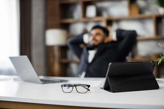 Focus on eye glasses, tablet and laptop. Happy relaxed caucasian young male rest in chair distracted from computer work, relieve negative emotions. Desktop background, copy space.