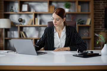 Young pretty businesswoman typing on laptop during work in office. Concentrated adult successful woman wearing official suit sitting at wooden desk, indoor.