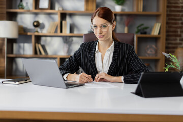 Young pretty female accountant working from modern office using laptop while holding a document and pen in her hands and sign new deal or writing report.