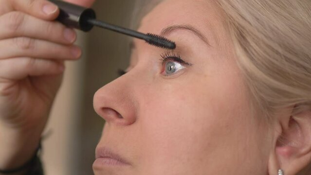close-up of an attractive caucasian blonde over 40 years old paints her eyelashes with mascara, selective focus. blonde mature in front of the mirror applying makeup