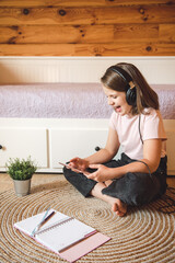 Cute smiling caucasian teen girl with black headphones playing game or having video chat using digital tablet and sitting at home on the floor next to bed.
