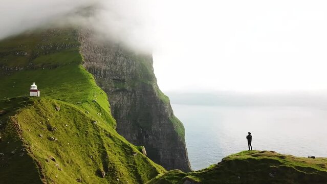 Aerial drone view of Kalsoy island with a man standing on a cliff, Faroe Islands. Amazing Faroese nature, green hills and beautiful rocky cliffs and mountain. Kallur lighthouse. Sunny day. 4k footage