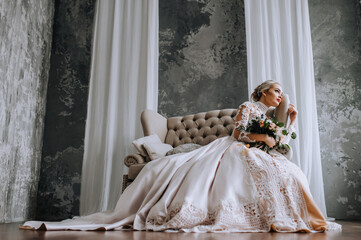 A beautiful blonde bride in a long lace dress sits on a sofa in the studio, indoors with a bouquet...