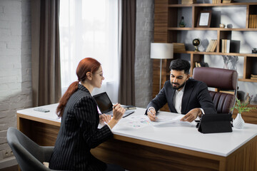 Handsome bearded businessman dealer showing papers with new agreement to his red hair businesswoman customer in a desktop, sitting at table in modern office.