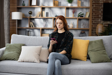 Obraz premium Attractive caucasian red hair woman sitting on couch and using modern smartphone indoors. Female freelancer taking break during working process at home.