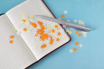 An open checkered notepad and a silver fountain pen lie on a blue background next to colorful confetti flowers. Concept of literary creativity, sketches, lyrics and poetry. Copy space