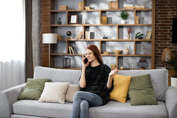 Fototapeta na wymiar Beautiful caucasian positive red hair woman sitting on couch and calling to her friend using smartphone indoors. Smiling female freelancer taking break during working process at home.