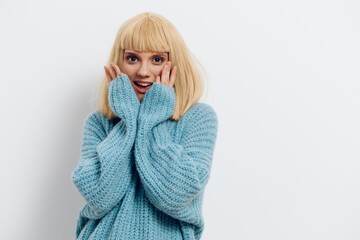 a close horizontal photo of a beautiful, sweet, pleasant woman in a stylish light blue sweater, smiling happily standing on a light background and holding her hands near her face