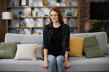 Obraz premium Happy red hair caucasian woman sit rest on comfortable couch in modern living room distance dreaming. Smiling young female renter or tenant relax on sofa at home, enjoy weekend.