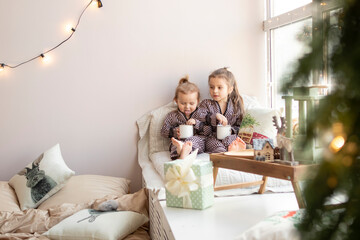 two sisters girls drink cocoa at home next to the window at christmas. new year concept