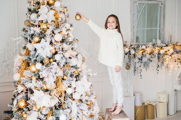Small adorable female child in white sweater and trousers holds toy for decoration, decorates New...