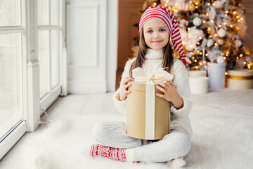 Happy little small child wears white knitted sweater holds gift sits in cozy room against New Year tree, feels comfort, glad to recieve Christmas present from parents. Childhood, holidays concept