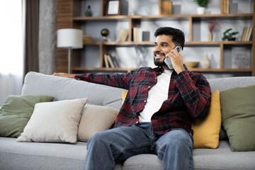 Fototapeta premium Handsome bearded positive man sitting on couch and calling to his friend using smartphone indoors. Smiling male freelancer taking break during working process at home.
