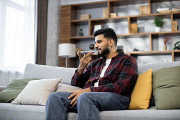 Attractive man in red checkered shirt sitting on couch and using modern smartphone indoors. Male freelancer taking break during working process at home.