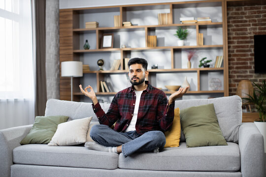 Calm of bearded young man doing yoga lotus pose to meditation and relax on couch during work online at home. Happy male sitting on sofa after hard work during break and deep breath in yoga posotion