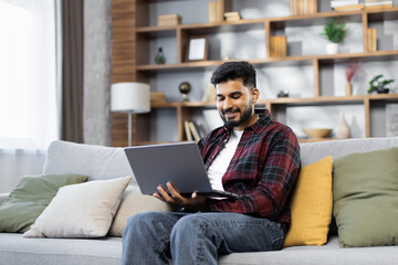 Bearded young man using laptop, sitting on couch at home, handsome guy having video call online in social network, having fun, watching movie, freelancer working on computer project