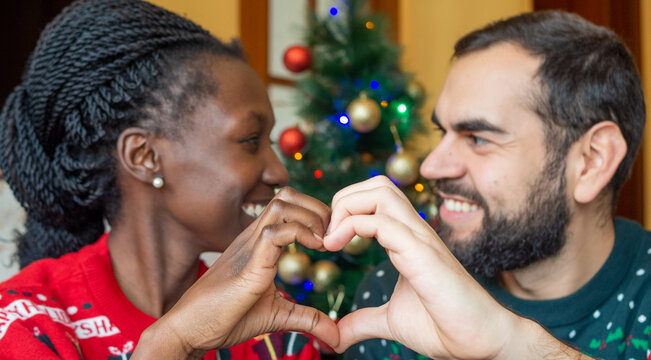 interracial couple enjoying festive season making a heart shape with their hands looking at each other in front of a Christmas tree while wearing christmas sweaters.