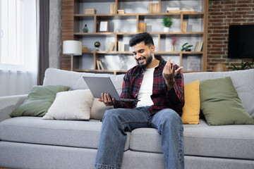 Obraz premium Young bearded businessman in checkered red shirt working on laptop computer, talking online while sitting on the comfortable sofa at home. Concept of remote work from home.