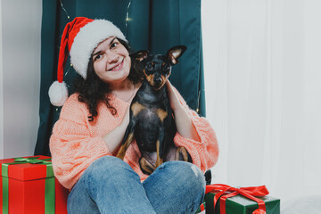 Portrait of young woman playing with funny dog miniature pinscher around Christmas ball and gift...