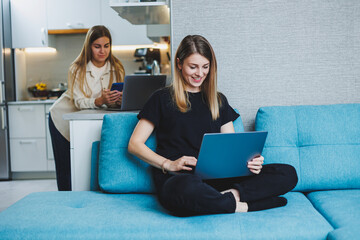 A young pretty woman is sitting on the sofa at home and working on a laptop, in the background another woman is talking on the phone in the kitchen. Two friends work at home.