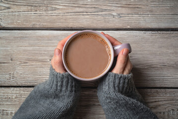 Female hands in a grey sweater holding a mug of hot chocolate on a rustic wooden background with a copy space