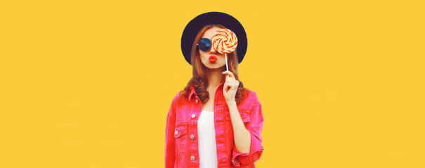 Portrait of happy young woman holding lollipop and blowing her lips sends air kiss wearing pink jacket, black round hat on yellow background