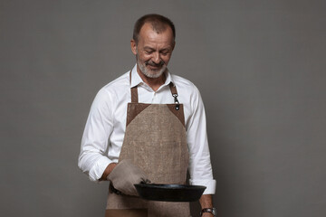 A mature man in a chef's apron holds a cast-iron frying pan with an oven mitt.