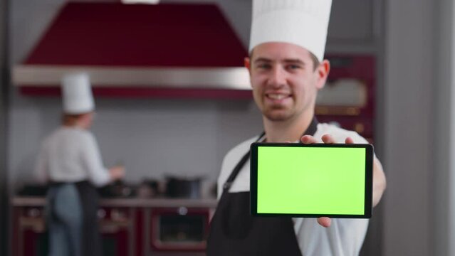 Chef holds smartphone wit a green screen in his hand and smiles standing in the kitchen. Realtime