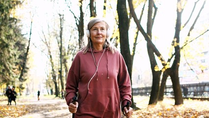 Happy active energetic mature woman hiking walking in urban autumn park using trekking poles...