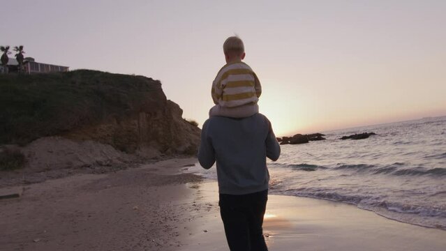 Little boy is sitting on father's neck, they having walk on he beach at sunset
