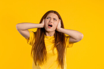 Fototapeta premium Annoyed ginger woman screaming with shut ears on yellow background. Lady block annoying sounds, music, shouts or noise. Rolling eyes.