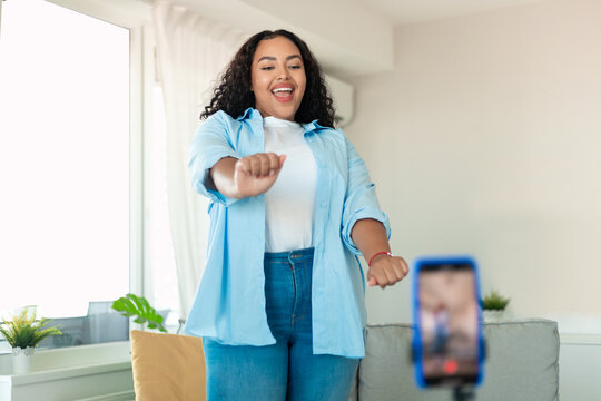 Cheerful black chubby woman dancing at cellphone camera filming video using phone on tripod at home