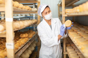 Focused Asian woman engaged in cheesemaking dressed in white uniform with cap, gloves and mask...