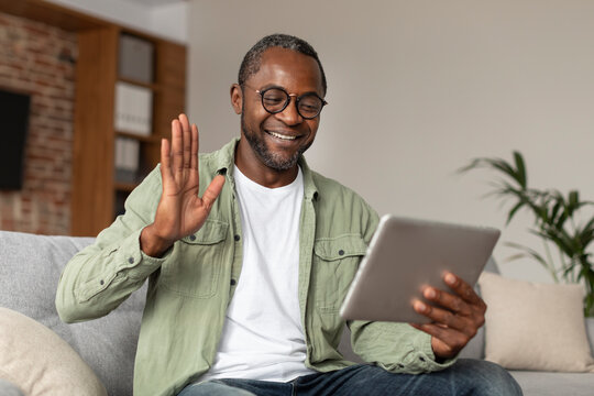 Glad happy middle aged black guy in glasses waving hand at digital tablet webcam, say hello, hi, make greeting gesture