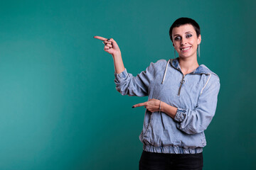 Smiling brunette woman with short hair pointing to side with forefinger advertising product while standing over isolated background. Female with positive facial expression showing promotion