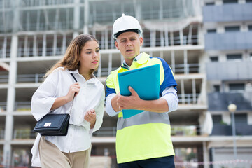Man engineer and a young woman customer discuss a construction project, standing on a construction site, holding a folder ..with documents