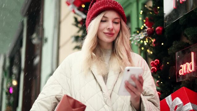 Charming blond young woman in red hat with present box hold smartphone scrolling social media texting and chatting with family or friend on snowy city street alone Preparation for Christmas New year