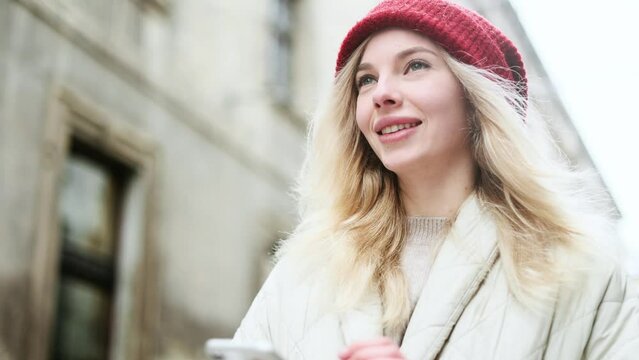 Portrait of charming smiling blond young woman with red hat hold smartphone scrolling social media texting browsing online on snowy winter street in city centre alone