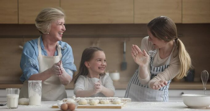 Multigenerational Women In Aprons Having Fun While Make Pastries, Little Girl Her Granny And Mom Clap Hands And Throw Flour While Prepare Dough For Homemade Pie. Family Recipe, Funny Pastime, Cookery