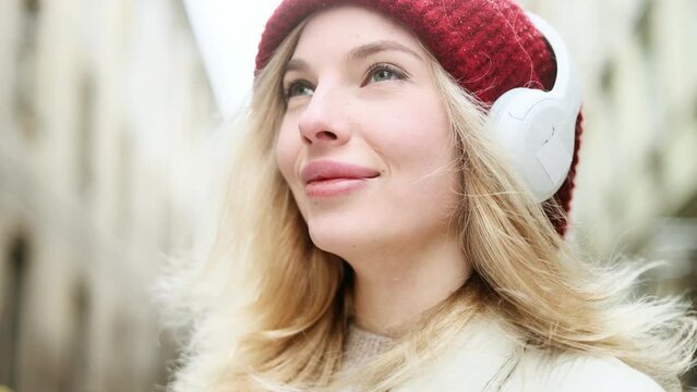 Close up portrait of charming smiling blond woman with red hat listening to music in headphones while standing on snowy winter street in city centre and inspired looking ahead alone