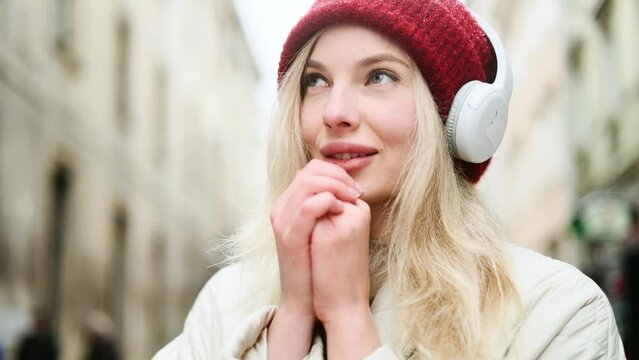 Close up portrait of charming blond young woman with red hat and headphones listening music while standing on snowy winter street in city centre and rubbing her hands to keep warm alone 
