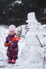 A four-year-old girl builds a snowman in the winter in the forest, holding a red shovel in her hand, surrounded by snowdrifts. Vertical orientation.