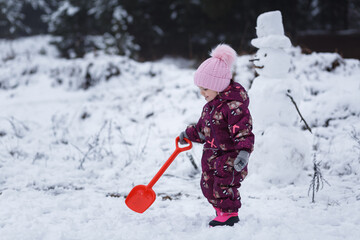 A four-year-old girl builds a snowman in the winter in the forest, around a snowdrift. Red spatula.