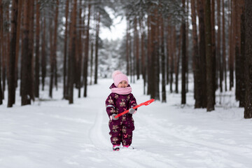 Fototapeta na wymiar A little girl stands in the middle of a snowy forest with a red shovel in her hands. Looks into the frame. Winter. Snowdrifts are white.
