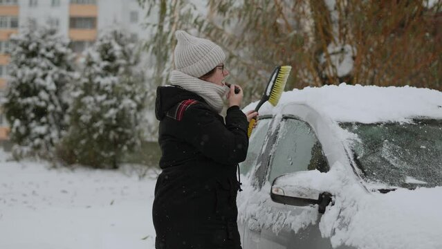 Pretty woman in warm clothes cleans snow from car and paints lips with lipstick. Lady is preparing car for driving in parking lot on frosty winter day. Concept do several things at once..