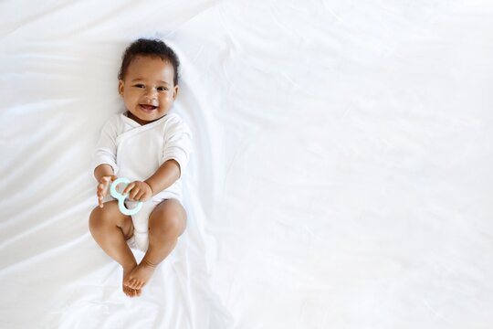 Childcare. Adorable Little Black Baby Lying On Bed With Teether In Hand