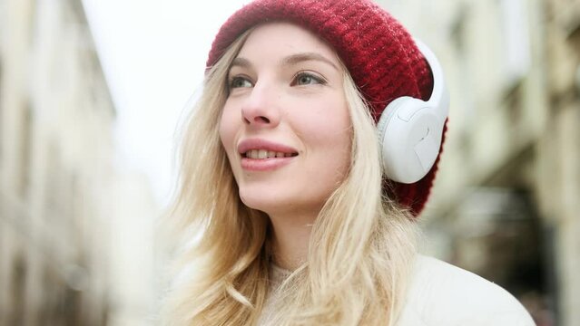 Close up portrait of charming smiling blond woman with red hat listening to music in headphones while standing on snowy winter street in city centre and inspired looking ahead alone